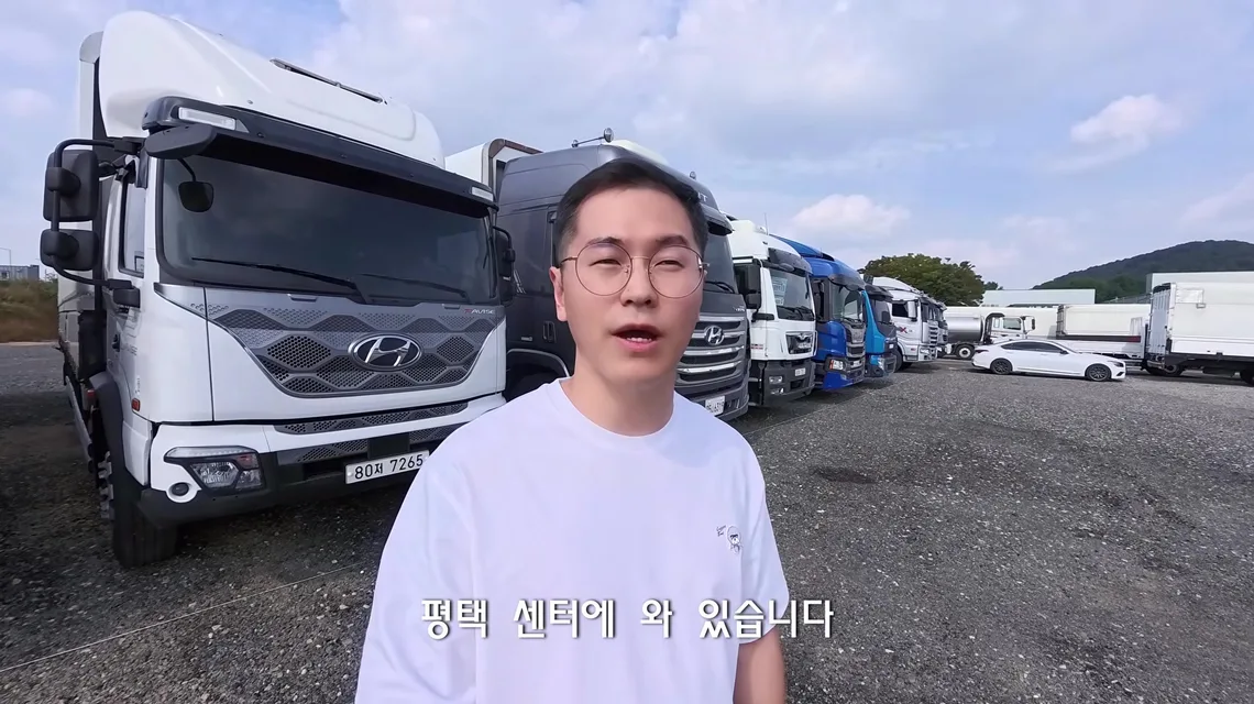 흰색 현대 엑시언트와 대형 트럭이 줄지어 선 아이트럭 평택센터 전시장에서 카메라를 보고 말하는 트럭커 태권v