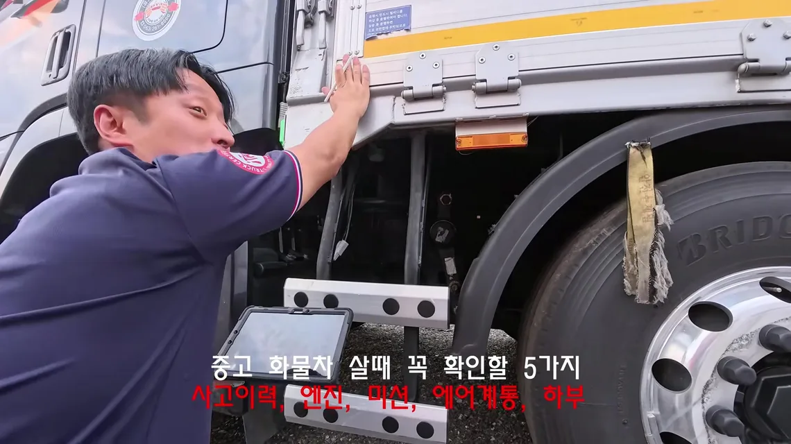 윙바디 적재함 옆 스텝과 타이어 앞에서 이동선 수석실장이 차체를 짚고 있고, 화면에 중고 화물차 살 때 꼭 확인할 5가지 사고이력 엔진 미션 에어계통 하부 자막이 나온 장면