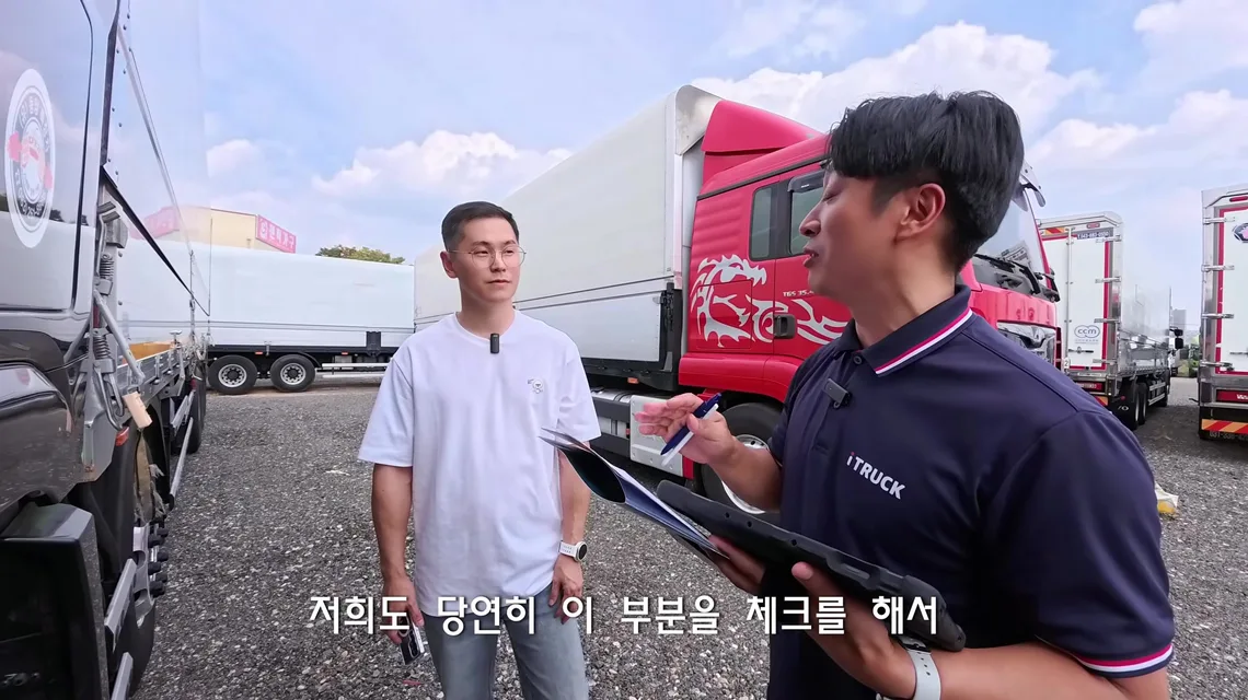 빨간 대형 트럭과 냉동탑차가 서 있는 전시장에서 이동선 수석실장이 태블릿과 서류를 들고 트럭커 태권v에게 보험사고 이력 조회를 설명하는 장면