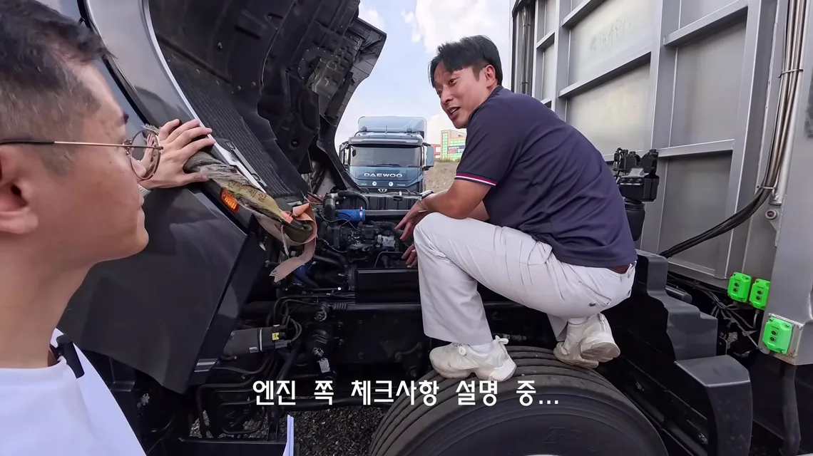 캡을 앞으로 젖혀 드러난 엑시언트 엔진룸 옆 타이어 위에 올라가 엔진 상부를 가리키며 설명하는 이동선 수석실장과 이를 들여다보는 트럭커 태권v