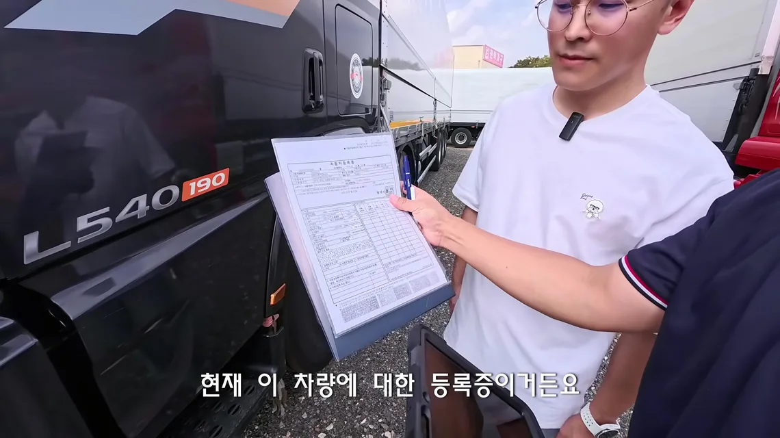 검은색 엑시언트 L540 캡 옆에서 이동선 수석실장이 클립보드에 끼운 자동차 등록증을 펜으로 짚으며 트럭커 태권v에게 설명하는 장면