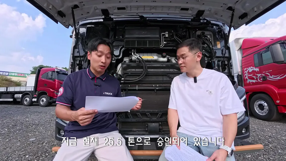 캡을 젖혀 엔진룸이 드러난 엑시언트 프로 540 앞에 앉아 서류를 보며 25.5톤 승인 제원을 설명하는 이동선 수석실장과 트럭커 태권v